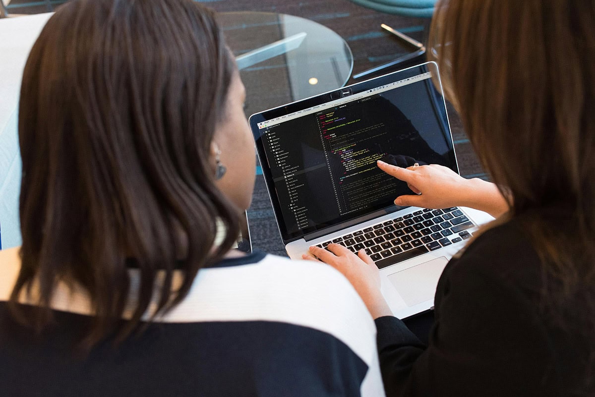 Two women pointing to and looking at code on laptop