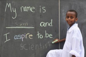 Young black boy writing "My name is same and I aspire to be a scientist" on a chalkboard