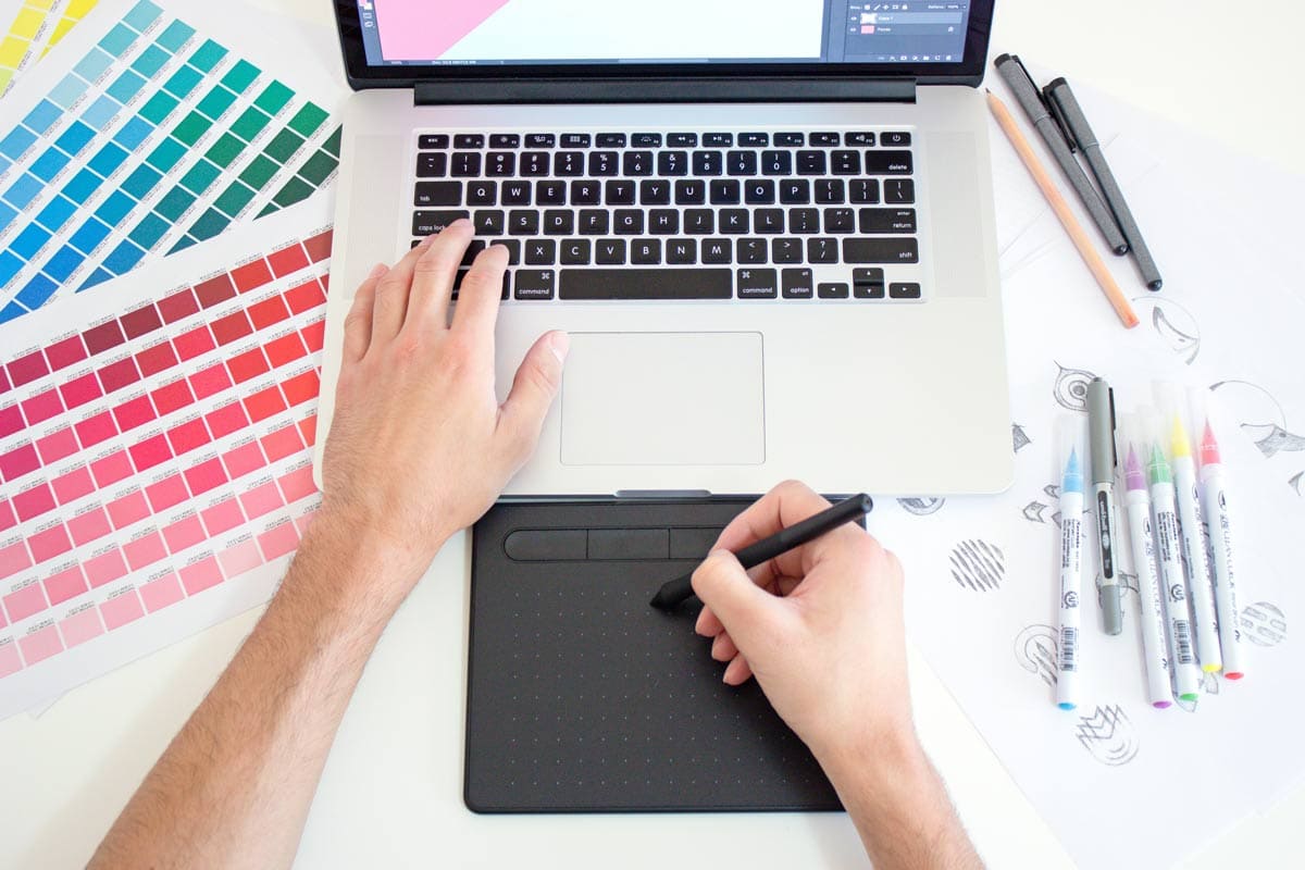 Overhead view of color palettes, pens, and doodles, with hand on laptop keyboard and other on digital drawing pad