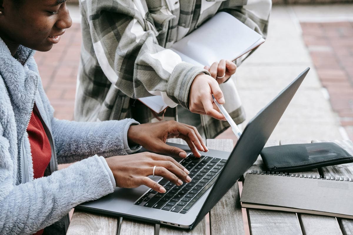 Students working together outside on a laptop, one girl with hands on the keyboard and the other pointing to the screen with a pen.