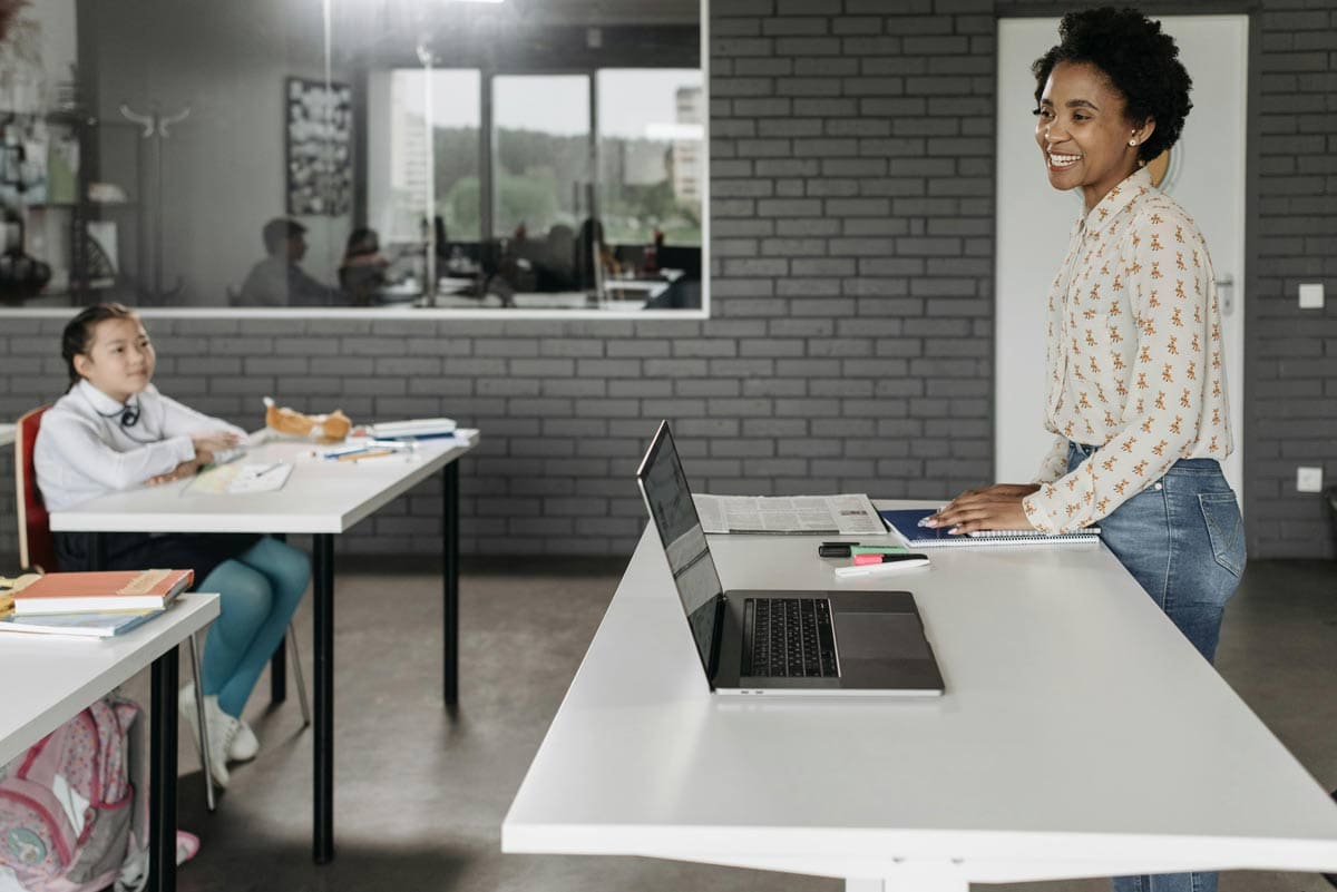 Smiling teacher in front of classroom with grey brick walls and laptop on white desk