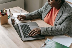 Teacher sitting at desk with left hand on lap top and right hand making notes on a pad of paper